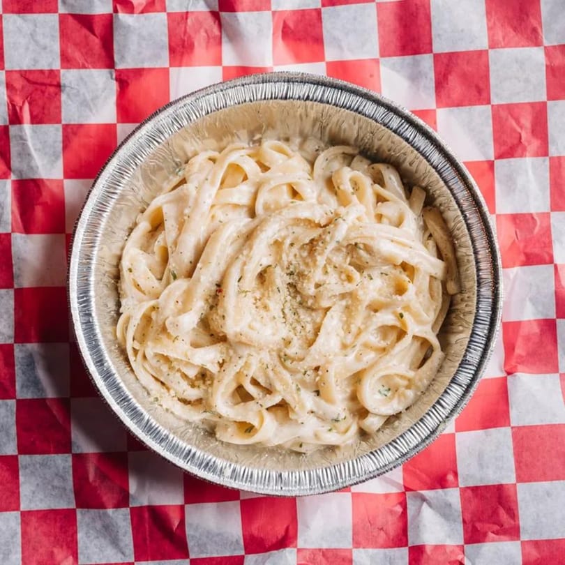 Creamy fettuccine alfredo with black pepper and parmesan in a foil container on red and white checkered paper.