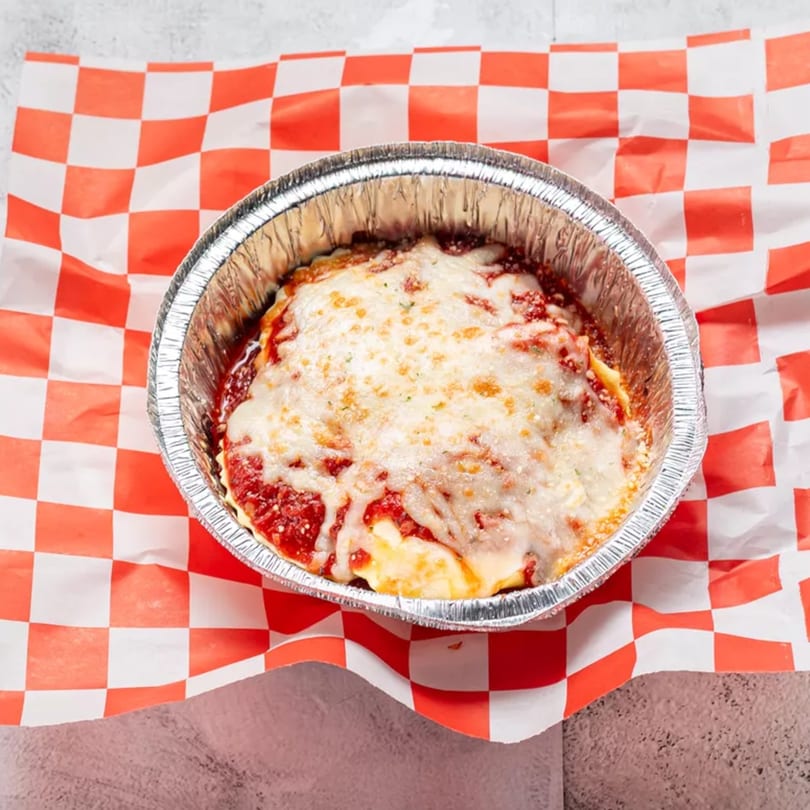 Baked lasagna in a foil container, topped with melted cheese and red sauce on checkered paper.