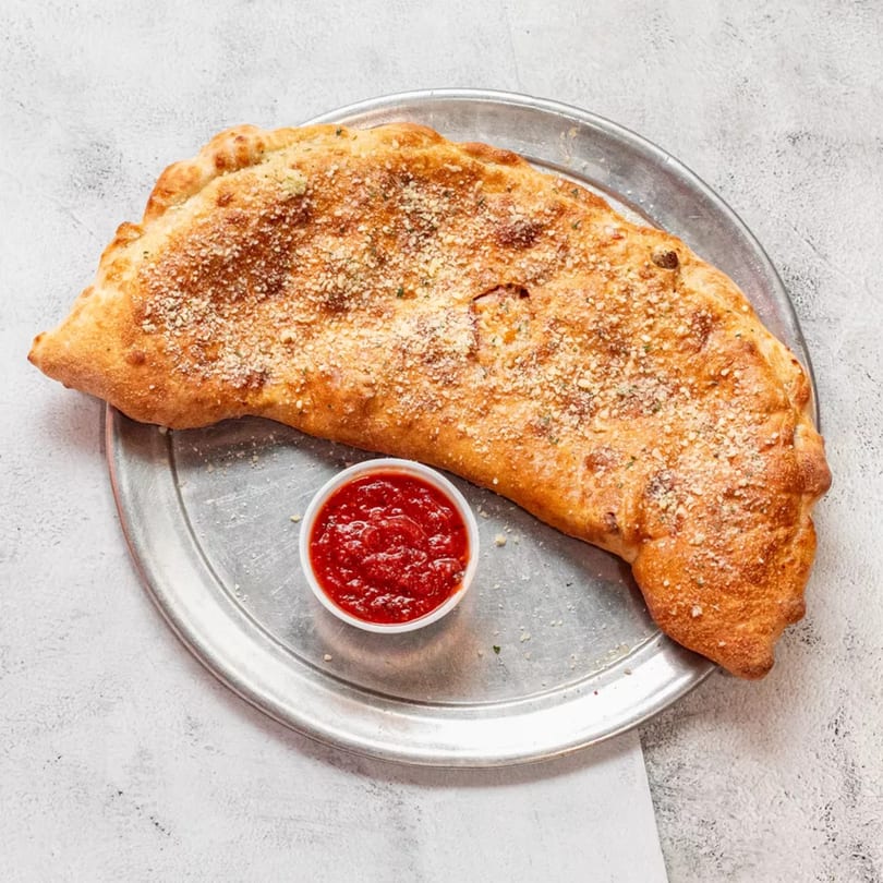 A golden-brown calzone sprinkled with parmesan cheese, served on a metal tray with a side of marinara sauce.