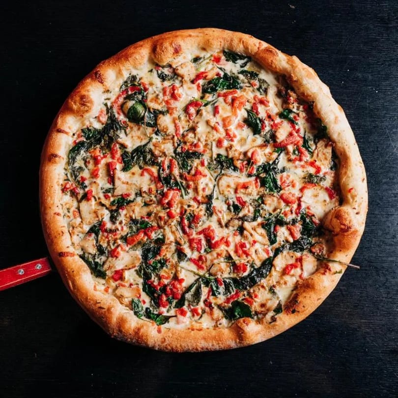 Round pizza with white sauce, chicken, spinach, and red peppers on a dark surface.
