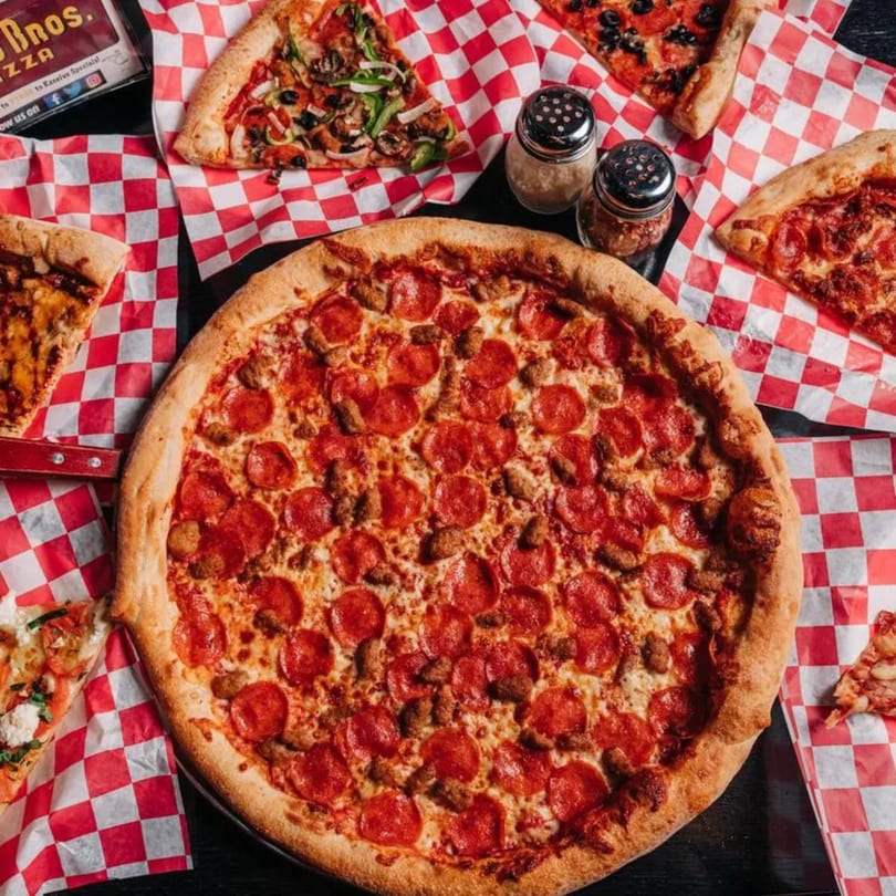 A large pepperoni and sausage pizza surrounded by various pizza slices on checkered paper.