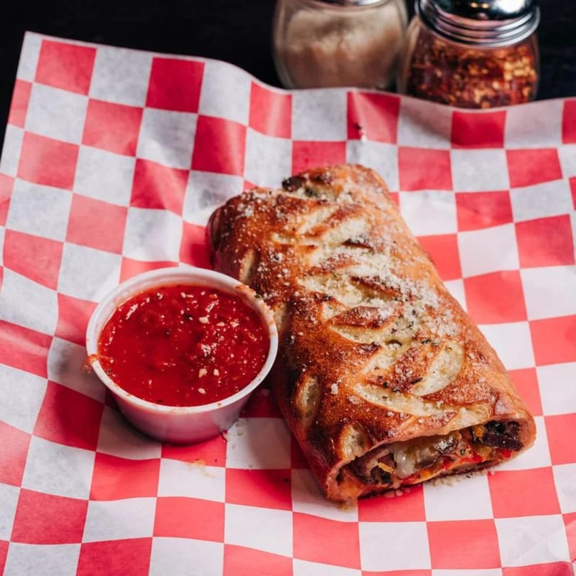 Golden-brown stromboli with parmesan cheese, served with a side of marinara sauce on checkered paper.