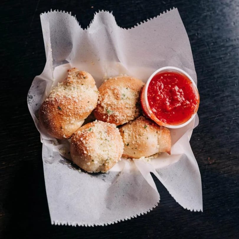 Garlic knots sprinkled with parmesan cheese, served in a paper-lined basket with a side of marinara sauce.