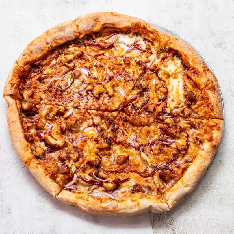A whole BBQ chicken pizza with red onions and melted cheese on a light background.
