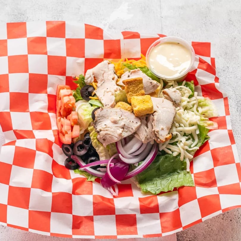 Chicken salad with lettuce, tomatoes, olives, onions, cheese, croutons, and dressing in a red and white checkered paper.