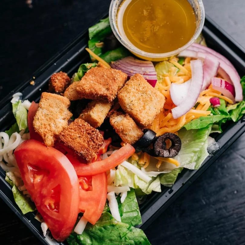 A fresh salad in a black plastic container with croutons, tomatoes, onions, olives, and dressing.